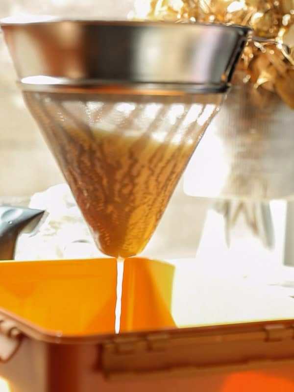 A liquid is being filtered through a cone-shaped mesh strainer into a container. The setup is in a bright, sunlit area, creating a warm, reflective ambiance.